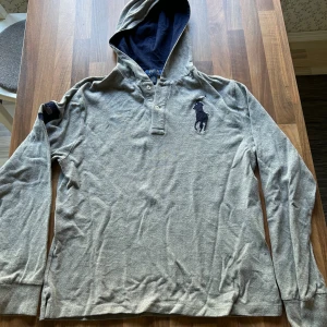 Grå hoodie från Polo Ralph Lauren - Grå hoodie från Polo Ralph Lauren med mörkblå broderad logga på bröstet och en stor mörkblå siffra 3 på ärmen. Tröjan har knappar vid halsen, huva med mörkblå insida och är tillverkad i mjuk bomull. Perfekt för en chill och sportig stil.