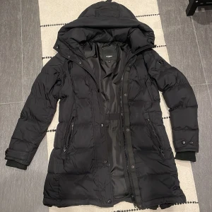 Svart lång pufferjacka Rockandblue - Svart lång pufferjacka från Rockandblue med huva, dragkedja och knappar framtill. Jackan har två sidofickor med dragkedja, ribbade muddar vid ärmslut och justerbara detaljer vid handlederna. Perfekt för kalla dagar med sin vadderade design och stilrena look. Jackan är hel men på höger armen så är det ett litet snitt men det syns inte har ni frågor så Ede bara att höra av er så kan jag skicka bild på just det:)