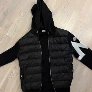 Svart Moncler Cardigan jacka  - Svart pufferjacka från Moncler med huva och dragkedja framtill. Jackan har stickade ärmar med vit zigzag-mönster och Moncler-logga på ena ärmen. Kroppen är quiltad och har två fickor med knappstängning. Jag har använt jackan i ungefär en månad. Säljer jackan eftersom jag inte har behov av den längre. Jackan är i storlek M men passar också bra om du har storlek S. Själv har jag storlek S och den passar perfekt. Jag kan frakta om det behövs(: jackan kommer men en moncler zip påse.