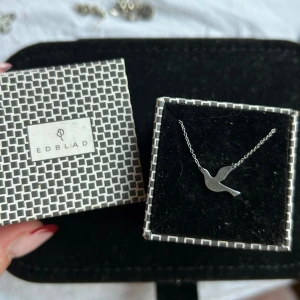 Edblad duva i stål halsband 42-45cm - Snyggt halsband från Edblad i silverfärgat rostfritt stål med en stilren duva som hänge. Kedjan är tunn och smidig, perfekt för att ge en minimalistisk touch till din look. Kommer i originalask med grafiskt mönster. Lilla Edblad berlocken vid knäppet har gått av men det är från Edblad