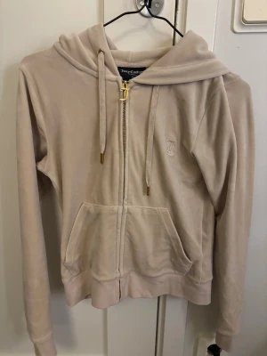 Beige velour  Juicy Couture XS - Snygg beige hoodie från Juicy Couture i mjuk velour med guldfärgad dragkedja och J-detalj. Tröjan har huva, snörning och fickor framtill samt broderad logga på bröstet. Perfekt för chill dagar och streetstyle.
