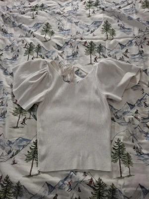 Vit ribbad blus med puffärm från ONLY - Supersöt vit blus från ONLY i ribbad bomull med puffiga korta ärmar och v-ringning. Toppen har en snygg knytning i ryggen som ger extra detalj. Perfekt för dig som gillar en feminin och trendig look med lite volym på axlarna.