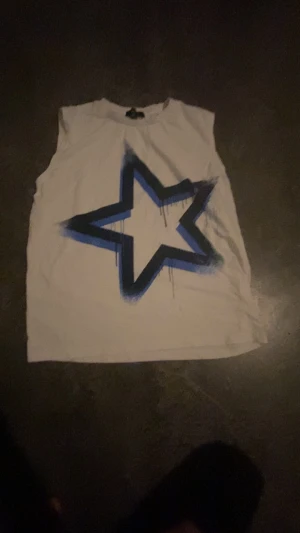 Vit ärmlös linnetopp med blå stjärna - Cool vit linnetopp från Oxide i storlek 158/164. Toppen är ärmlös och har en stor blå och svart stjärna med spray-effekt på framsidan. Perfekt för dig som gillar streetstyle och vill sticka ut med ett grafiskt tryck.