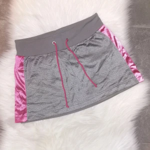 Grå och rosa sportig kortkjol från H&M, storlek XS - Sportig kortkjol i grått med glansiga rosa paneler på sidorna och rosa snörning i midjan. Kjolen har bred resår upptill och är tillverkad i ett syntetmaterial med mesh på utsidan. Storlek XS, köpt på H&M. Är i mycket fint skick. 