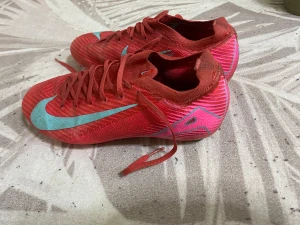 Nike röda fotbollsskor Flyknit - Säljer ett par röda Nike fotbollsskor med turkosa detaljer och logga. Skorna har snörning, mönstrad ovandel i syntet och stickat material (Flyknit) samt dobbar i grönt och rosa undertill. Perfekta för dig som vill sticka ut på planen. Dem är pros