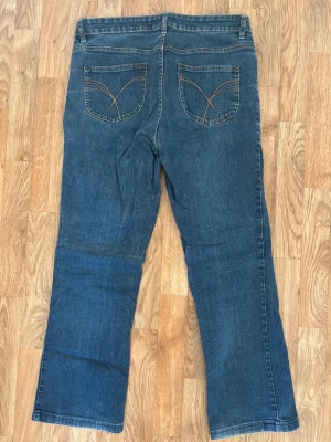 Blå bootcut jeans från Jeanswear, stl 42L - Snygga blå jeans från Jeanswear i storlek 42 Long med klassisk bootcut passform. Jeansen har kontrastsömmar, fem fickor och coola broderade detaljer på bakfickorna. Perfekt för dig som gillar lite bredare ben och tidlös denimstil.