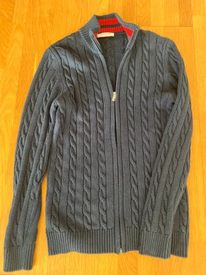 Mörkblå stickad kofta från Felix Hardy - Säljer en mörkblå stickad kofta från Felix Hardy med dragkedja framtill och snyggt kabelstickat mönster. Insidan av kragen har coola röda och mörkblå ränder som ger extra stilpoäng. Perfekt för dig som gillar klassisk men modern look.