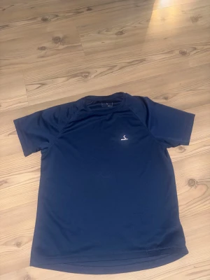 Blå tränings-tshirt från Energetics - Säljer en marinblå t-shirt från Energetics i storlek L, perfekt för träning. Materialet är lätt och syntetiskt med DRY PLUS-teknologi som håller dig torr. T-shirten har korta ärmar och en diskret logga på bröstet. Enkel och sportig design.