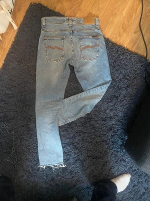 Ljusblå Nudie Jeans W29 L30 - Säljer ett par ljusblå jeans från Nudie Jeans med klassisk femficksdesign och snygga broderade detaljer på bakfickorna. Jeansen har en rak passform och fransade benslut för en cool, avslappnad vibe. Perfekt för dig som gillar streetstyle. 