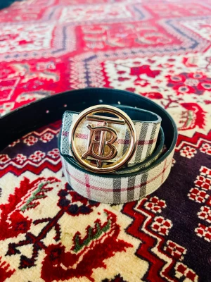 Burberry rutigt bälte med guldspänne - Snyggt bälte från Burberry med klassiskt rutigt mönster i beige, svart och rött. Bältet har ett stort, rundat guldspänne med Burberrys ikoniska TB-logo. Utsidan är i präglat skinn med mönster och insidan är svart. Perfekt för att lyfta din outfit med en lyxig touch.