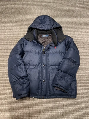 Mörkblå dunjacka från Polo Ralph Lauren - Säljer en fet mörkblå dunjacka från Polo Ralph Lauren med huva och röd logga på bröstet. Jackan har både dragkedja och knappar framtill, samt ribbade muddar vid ärmslut. Perfekt för kalla dagar och har en schysst oversized vibe.