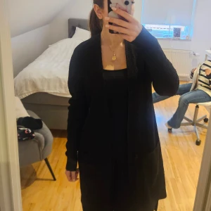 Svart stickad cardigan jacka med bälte - Säljer en svart, lång och mysig stickad cardigan jacka med bälte i midjan. Jackan har långa ärmar, två stora fickor framtill och en öppen design som kan knytas med bältet. Perfekt för lager-på-lager och chill stil.
