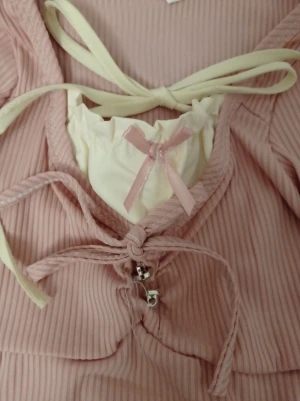 Rosa ribbad tjöja med rosett - Supersöt rosa ribbad tröja med långa ärmar och vita knytband. Framtill finns en dekorativ rosa rosett och små silverfärgade detaljer. Tröjan har en romantisk vibe med volangkant och v-ringning.obs denna tröja är tight så skulle säga den passar någon som är xs eller xss 