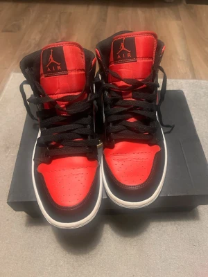 Nike Air Jordan 1 Mid Black/Red/White - Säljer ett par Nike Air Jordan 1 Mid i färgerna svart, röd och vit. Klassisk high-top siluett med svart snörning, röd tåbox och detaljer, vit sula och sidopaneler samt ikoniska Jordan Wings-loggan på sidan. Skorna är tillverkade i skinn och har en riktigt clean streetstil.