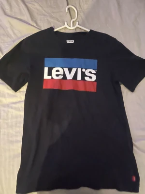 Svart Levi's t-shirt med logga - Svart t-shirt från Levi's med stor, klassisk logga i vitt, blått och rött på bröstet. Modellen är kortärmad och har rund hals. Materialet är mjuk bomull som känns skön mot huden. Perfekt för dig som gillar streetwear och ikoniska märken.
