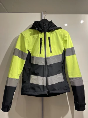 Gul och svart vindjacka med reflex - Grym vindjacka i neongult och svart med breda reflexränder och huva. Jackan har dragkedja framtill, två bröstfickor med dragkedja och justerbara ärmslut. Den är vattentät och sitter jätte fint, passar till det mesta både yrkesmässigt men även till fritiden💚