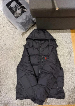 Polo jacka  - Svart pufferjacka från Polo Ralph Lauren med huva och dragkedja framtill. Jackan har det klassiska röda Polo-logot broderat på bröstet och två dragkedjefickor. Materialet är glansigt och jackan är vadderad för extra värme. Perfekt för kalla dagar.