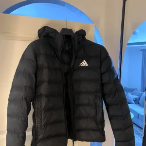 Svart pufferjacka från Adidas - Svart pufferjacka från Adidas med huva och dragkedja. Jackan har en quiltad design och Adidas-logga på bröstet. Tillverkad i syntetmaterial, perfekt för kalla dagar. Klassisk passform och långärmad modell.