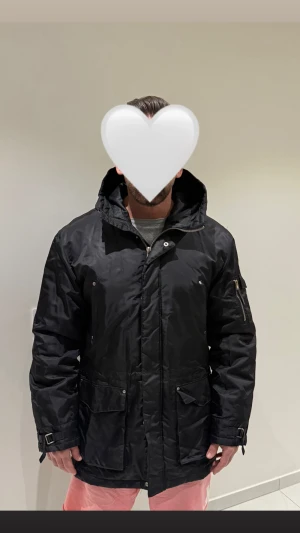 Svart parkajacka från River Island - Svart parkajacka från River Island med huva, dragkedja och knappar framtill. Jackan har flera stora fickor, justerbara spännen vid ärmslut och innerficka. Materialet är syntet och jackan är quiltad på insidan för extra värme.