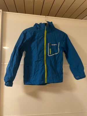 Everest Barn Regnjacka  - Everest barnjacka i storlek 134/140, blå färg med gröna detaljer, sido- samt bröstficka.   Jackan är unisex och har en avtagbar huva samt dragkedja. Materialet är anpassat för barn och passar för höst, vinter och vår✨