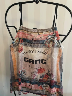 Blommig spetslinne från GANG - Säljer ett färgglatt linne från GANG med tunna axelband, blommigt mönster och spetsdetaljer. Toppen har tryckt text framtill och mix av rosa, blå, beige och orange toner. Perfekt för dig som gillar unika och lekfulla plagg med mycket detaljer.