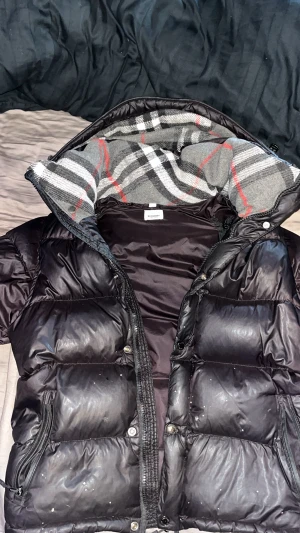 Svart pufferjacka från Burberry - Säljer en svart pufferjacka från Burberry London England med klassiskt rutigt foder i grått, vitt, svart och rött. Jackan har dragkedja, knappar och flera fickor med dragkedja. Oversized look och riktigt varm, perfekt för vintern, små hål på båda sidorna vid fickorna annars inget
