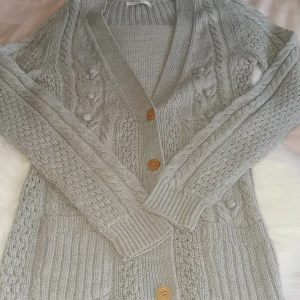 Only grå kofta, storlek XS - Supermysig ljusgrå stickad kofta från ONLY med snygga flätstickade detaljer och knappar. Koftan har långa ärmar, två fickor framtill och bred ribbad kant. Tröjn är i använd skick, har två lösa "bollar" där sömmen har lossnat lite vilket går enkelt att sy fast :), se bild . Storlek XS. Från Only. 