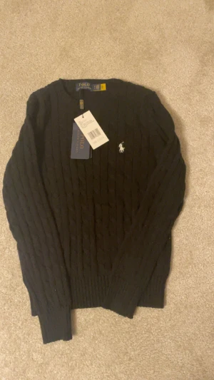 Svart kabelstickad tröja Polo Ralph Lauren - Svart kabelstickad tröja från Polo Ralph Lauren med klassisk rund hals och vit broderad logga på bröstet. Tröjan har långa ärmar och ribbade muddar vid ärmslut och nederkant. Perfekt för dig som gillar stilrena och tidlösa plagg.