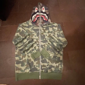Camouflage hoodie med hajmotiv - Säljer en grön camouflage hoodie från BAPE med hel dragkedja och hajmotiv på huvan. Tröjan har ribbade muddar, fickor fram och coolt tryck som sticker ut. Perfekt för dig som vill ha streetwear med edge.