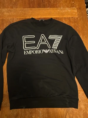 Svart EA7 Emporio Armani sweatshirt - Svart sweatshirt från EA7 Emporio Armani med stor vit logotyp framtill. Tröjan har rund hals och långa ärmar, tillverkad i mjukt material som känns skönt mot huden. Perfekt för dig som gillar stilrena och exklusiva märken.