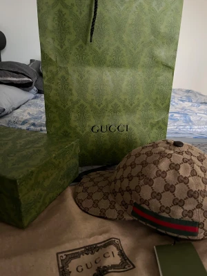 Gucci beige keps med GG-mönster - Snygg beige keps från Gucci med klassiskt GG-monogram över hela kepsen och en grön-röd rand på sidan. Justerbar rem baktill i brunt skinn och insida i mörkbrunt tyg. Perfekt för dig som vill ha en lyxig och trendig accessoar.