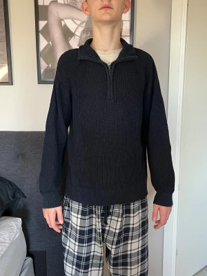 Svart stickad tröja med half zip - Svart stickad tröja med ribbad struktur och halv dragkedja vid kragen. Modellen är långärmad och har en klassisk, lite loose passform. Perfekt för kyliga dagar och enkel att matcha med jeans eller mjukisbyxor. Storlek L i barn från kidsbrandstore