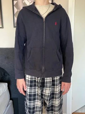 Mörkblå hoodie från Polo Ralph Lauren - Snygg mörkblå hoodie från Polo Ralph Lauren i storlek XL. Tröjan har dragkedja, huva och två stora fickor framtill. Klassisk röd Polo-logga på bröstet och mjukt bomullsmaterial som är skönt att ha på sig. Perfekt för chill dagar och enkel att matcha med jeans eller mjukis.