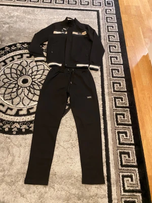 Svart bomberjacka från BOSS, strl S - Svart bomberjacka från BOSS med beige och vita ränder på krage och ärmslut. Jackan har dragkedja, ribbade muddar och BOSS-logga på bröstet. Tillverkad i mjuk bomullsmix, perfekt för en clean och sportig look.