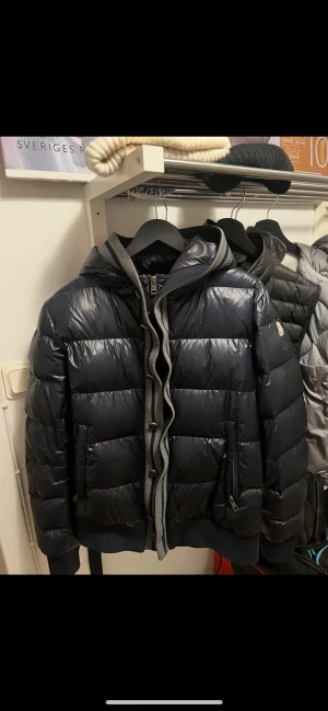 Svart Moncler pufferjacka med huva - Snygg svart pufferjacka från Moncler med glansig finish, huva och ribbade muddar. Jackan har dragkedja framtill, gråmelerade stickade detaljer och Moncler-logga på ärmen. Perfekt för kalla dagar när du vill hålla stilen. Köpt på kasperheats för 6500kr kemtvättad för 1 månadsen