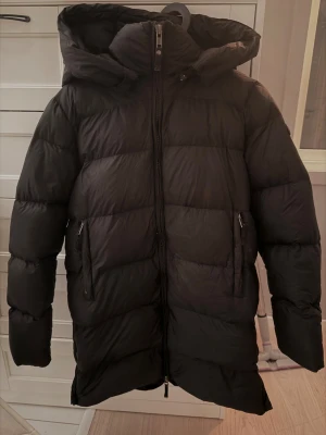 Svart dunjacka från Cedrico med huva - Säljer en svart dunjacka från Cedrico i storlek S. Jackan har en stor och mysig huva, två sidofickor med dragkedja och quiltad design. Perfekt för kalla dagar och har en stilren look med logotyp på ärmen. Jackan blev tyvärr aldrig använd så den är som ny 
