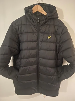 Svart pufferjacka från Lyle & Scott - Svart pufferjacka från Lyle & Scott med huva och dragkedja framtill. Jackan har det klassiska gula logobroderiet på bröstet och ribbade muddar vid ärmslut och nederkant. Perfekt för kyliga dagar och har en clean, sportig look.