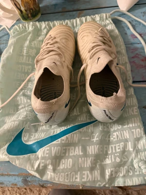 Nike Phantom GX fotbollsskor vita Elite - Nike Phantom GX fotbollsskor i ljus beige/vit med blå detaljer. Skorna har snörning och en rund tå, tillverkade i syntetmaterial med textilinslag. Sulan är designad för optimalt grepp på planen. Säljes med original Nike-påse. INGA HÅL ELLER TRASIG NÅGONSTANS. Bara lite smutsig.
