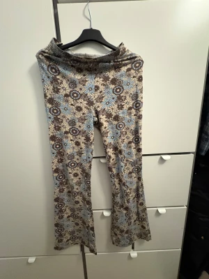 Blommiga bootcut byxor från Bik Bok - Supersnygga bootcut byxor från Bik Bok i storlek S. Byxorna har ett beige och brunt blommönster med ljusblå detaljer. De är stretchiga och har en skön passform med utsvängda ben. Perfekta för dig som gillar retro vibes och vill sticka ut lite extra.