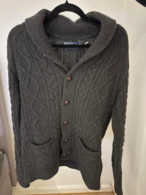 Mörkgrå stickad kofta från Polo Ralph Lauren - Mysig mörkgrå stickad kofta från Polo Ralph Lauren med grov kabelstickning, stora framfickor och knappar framtill. Koftan har en bred krage och långa ärmar, perfekt för kyliga dagar. De är en ull o kashimir blandning. Passar någon som är cirka 180cm lång. Använd ett fåtal gånger ca 3, så är som ny. Hör av er vid minsta fundering eller fråga.