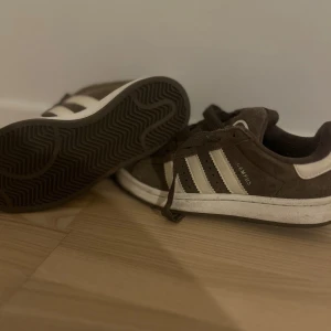 Adidas Campus bruna sneakers - Säljer ett par Adidas Campus sneakers i mörkbrunt mocka med vita ränder på sidorna och vit sula. Har endast använt de några gånger med försiktighet och de är i perfekt skick.
