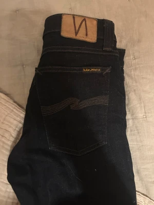 Mörkblå skinny jeans från Nudie Jeans - Snygga mörkblå skinny jeans från Nudie Jeans med klassisk femficksdesign och kontrastsömmar. Modellen har normal passform i midjan och smal siluett hela vägen ner. Materialet är slitstarkt jeans och detaljerna inkluderar kopparfärgade nitar och logopatch bak.