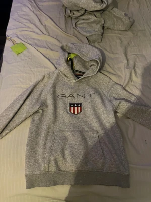 Grå hoodie från GANT med emblem 158/164 - Snygg grå hoodie från GANT med broderat emblem och logga på bröstet. Tröjan har huva, lång ärm och en stor magficka framtill. Perfekt för dig som gillar klassisk college-stil och vill ha något bekvämt och stilrent.