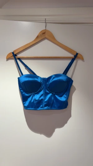 Blå satin korsett-topp med dragkedja - Snygg blå korsett-topp i glansigt satinmaterial med justerbara axelband och markerade kupor. Toppen har en dragkedja bak för enkel på- och avtagning. Perfekt för att skapa en trendig och edgy look.