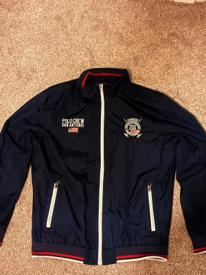 Marinblå vindjacka Vinson Polo Club - Snygg marinblå vindjacka från Vinson Polo Club med broderade detaljer och amerikansk flagga. Jackan har vita dragkedjor, ribbade muddar med röd och vit rand, samt brodyr på bröstet. Perfekt för dig som gillar sportig och stilren look.