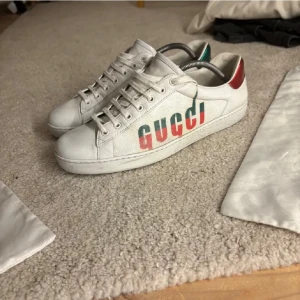 Vita sneakers från Gucci - Snygga vita sneakers från Gucci med klassisk logga i rött och grönt på sidan. Skorna har snörning och detaljer i rött och grönt på hälen. Perfekta för en stilren look. Kvitto finns vid förfrågan 