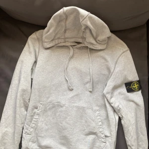 Grå hoodie från Stone Island - Säljer en stilren grå hoodie från Stone Island med klassisk patch på ärmen. Tröjan har huva med snörning, magficka och är tillverkad i mjuk bomull.  Priset är 1500kr men kan diskuteras.                Nypris är 3500kr