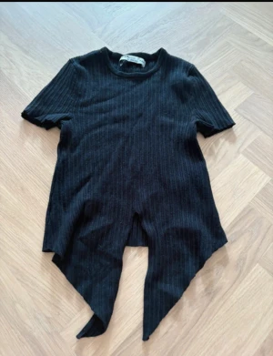 Svart ribbad topp från FB Sister - Svart ribbad topp från FB Sister med korta ärmar och unik asymmetrisk nederkant. Toppen är croppad och har en tight passform som framhäver siluetten. Perfekt för dig som gillar edgy och trendiga detaljer.