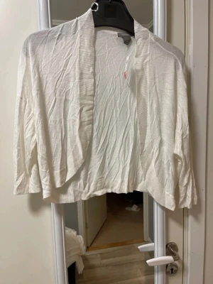 Vit kofta från Lindex - Säljer en vit, tunn och öppen kofta från Lindex med ribbade detaljer längs kragen och nederkant. Koftan har lång ärm och är gjord i mjuk bomull, perfekt att slänga över en topp när det är lite kyligt ute.