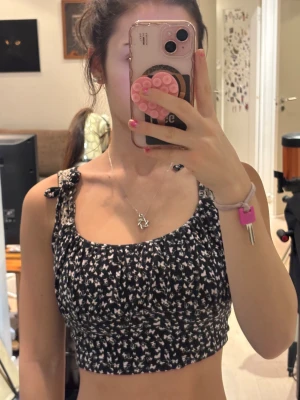 Svart topp med fjärilsmönster och knyt - Säljer en svart croppad topp med vita fjärilar och små rosetter i mönstret. Toppen har smala axelband som knyts på axlarna.